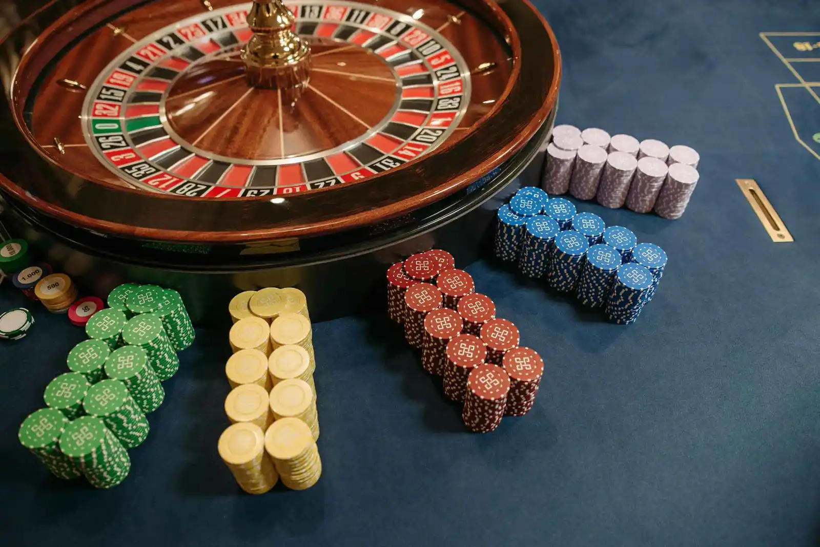 Rueda de ruleta fotografiada de costado sobre un paño oscuro, con torres de fichas de colores al lado.